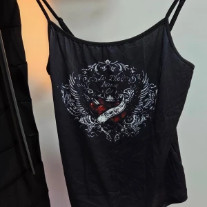 Svart linne med tryck och tunna band, Stl M - Säljer ett svart linne med tunna axelband och ett detaljerat tryck framtill med hjärta, vingar och texten 'In The Hands Of'. Toppen har en cool vibe och är perfekt för dig som gillar edgy stil. Materialet känns mjukt och lätt.