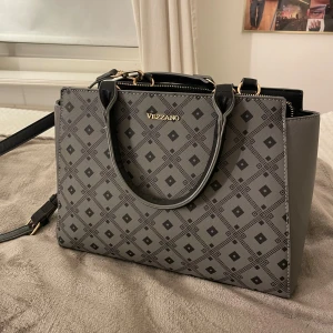 Mönstrad handväska från Vezzano - Snygg handväska från Vezzano i grått och svart med geometriskt mönster. Väskan har både handtag och axelrem, guldfärgade detaljer och flera fack med dragkedja. 
