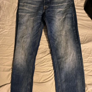 Blå tapered jeans från Jack & Jones - Snygga blå jeans från Jack & Jones i modellen Tapered/Mike. Jeansen har en klassisk femficksdesign, ljusa slitningar framtill och smalare ben som ger en modern look. Tillverkade i mjukt denimtyg med kontrastsömmar och normal passform.