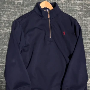 Marinblå half zip tröja Polo Ralph Lauren - Snygg marinblå långärmad tröja från Polo Ralph Lauren med half zip och klassisk röd logobrodyr på bröstet. Tröjan har en hög krage och är gjord i mjuk bomull med fleece på insidan. Perfekt för dig som gillar stilren och sportig look.