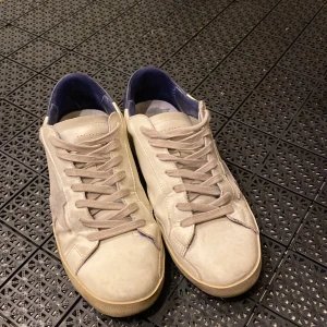 Golden Goose vita sneakers med blå detaljer - Säljer ett par snygga Golden Goose sneakers i vitt skinn med beige mockadetaljer och en stjärna på sidan. Skorna har blå hälparti, beige snörning och platt gummisula. Insidan är fodrad i blått och sulan har ett klassiskt fiskbensmönster. Perfekt för dig som gillar streetstyle.