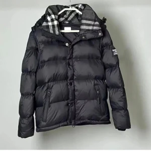 Svart dunjacka från Burberry - Svart dunjacka från Burberry med huva och dragkedja samt knappar framtill. Jackan har ett klassiskt rutigt foder i huvan och logotyp på ärmen. Två fickor med dragkedja och justerbar huva. Perfekt för kalla dagar.