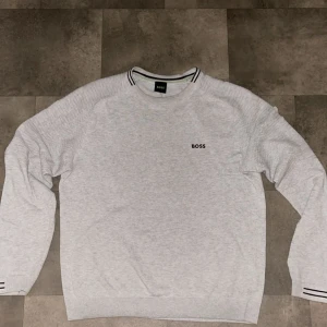 Ljusgrå sweatshirt från BOSS - Snygg ljusgrå sweatshirt från BOSS med svart logga på bröstet. Tröjan har rund halsringning med svarta och gröna detaljer samt ribbade muddar med svarta ränder vid ärmslut. Perfekt för en clean och stilren look.