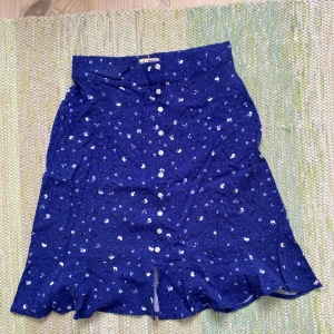 Blå blommig kjol från Holly & Whyte - Säljer en blå kortkjol från Holly & Whyte med små vita blommor, volangkant och knappar framtill. Kjolen har en söt och feminin stil, perfekt för dig som gillar detaljer och mönster. Elastisk midja för skön passform.