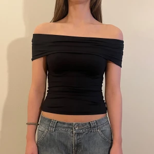Svart offshoulder topp - Säljer en svart offshoulder topp med tight passform. Toppen är croppad och har en bred kant som vilar över axlarna. Perfekt för dig som gillar en stilren och enkel look. Materialet är stretchigt och mjukt, vilket gör den bekväm att bära. Modellen är 171 cm lång.