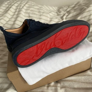 Mörkblå mocka sneakers Louboutin - Äkta & Snygga mörkblå sneakers från Christian Louboutin i mjuk mocka med klassisk röd sula och rund tå. Skorna har snörning och diskreta detaljer, samt beige innersida i skinn. Perfekt för dig som vill ha en lyxig och stilren look med en ikonisk touch.