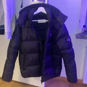 Svart pufferjacka från Calvin Klein - Säljer en svart pufferjacka från Calvin Klein med stor logga på ärmen. Jackan har huva, dragkedja framtill och två sidofickor. Perfekt för kalla dagar och riktigt snygg till streetwear-stilen.