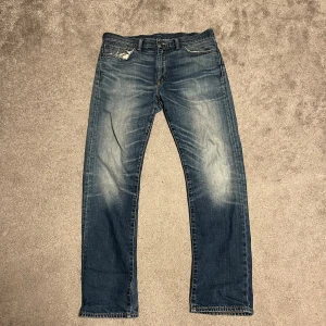 Levi's 504 blå jeans straight fit - Klassiska Levi's 504 jeans i blå tvätt med slitningar och ljusa partier framtill. Rak passform med normal midja och fem fickor. Jeansen har en liten reva vid myntfickan och tydliga kontrastsömmar. Perfekta för en avslappnad stil.
