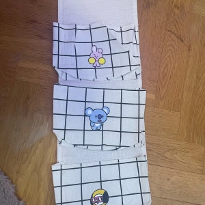 Rutiga förvaringsväskor med BT21 motiv - Tre praktiska förvaringsväskor i beige tyg med svart rutmönster och söta BT21 figurer på varje ficka som kan hängas upp på väggen. Perfekt för att organisera småsaker i rummet eller på skrivbordet. Väskorna har en enkel och stilren design med roliga karaktärer.