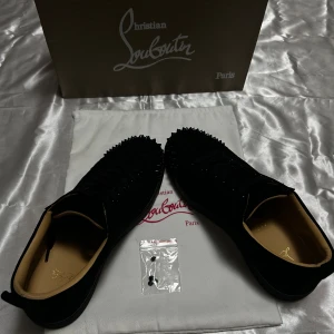 Christian Louboutin svarta sneakers med nitar - Svarta sneakers från Christian Louboutin i mocka med coola svarta nitar över hela tån och sidorna. Skorna har klassisk röd sula, snörning och en låg profil. Kommer med originalkartong och dustbag. Perfekta för dig som vill sticka ut med lyxig streetstyle.