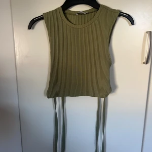 Olivgrön ribbad backless topp från Zara - Säljer en olivgrön ribbad linnetopp från Zara med croppad passform och knytband som man knyter bak. Toppen är ärmlös och har en backless look. Perfekt för både vardag och fest.