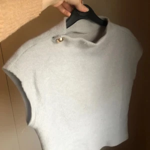 Grå stickad väst med guldfärgad knapp - Snygg grå stickad väst med hög hals och en guldfärgad knapp vid axeln. Västen har ribbad struktur och är ärmlös, vilket ger en clean och modern look. 