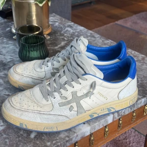 Premiata sneakers i vitt och blått, strl 42 - Säljer ett par Premiata sneakers i vitt skinn med blå detaljer och grå snörning. Snyggt crackle-mönster på ovandelen och texten BSKT/CLAY på hälen. Platt sula med grafiska tryck och perforerad tå. Perfekt för dig som gillar unik design och streetstyle.