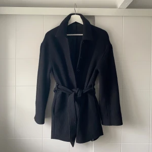Svart kappa med bälte  - Snygg svart Filippa K kappa med knappar framtill och ett matchande bälte i midjan. Jackan har en rak passform som ger en chill vibe men kan dras åt i midjan så att den blir mer figuranpassad. Köpt för 3 år sen men har inte används mycket pga att jag har liknande kappor. Storleken är i 36 men passar perfekt för mig som vanligtvis bär storlek 34/xs