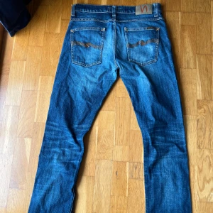 Blå jeans från Nudie Jeans, W32 L34 - Säljer ett par klassiska blå jeans från Nudie Jeans med snygga slitningar och kontrastsömmar. Modellen har raka ben, fem fickor och ikoniska broderade bakfickor. Perfekt passform och cool tvätt som funkar till allt. Model Grim Tim 
