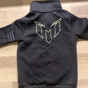 Svart Messi hoodie från Adidas - Säljer en svart quiltad hoodie från Adidas med Messi-logga på bröstet och ryggen. Tröjan har huva, half zip, magficka och långa ärmar. Snygga detaljer med vita dragkedjor och diskret Adidas-logga på ärmen. Perfekt för fotbollsfans!