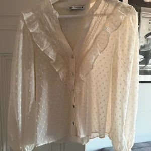Vit volangblus från Zara - Vit blus från Zara med volangdetaljer längs v-ringningen och knappar framtill. Tunt, lätt material med diskret prickigt mönster och långa ärmar med smock och volang vid ärmslut. 