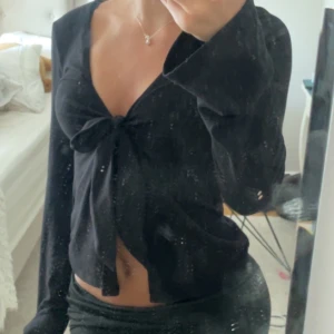 Svart långärmad knytblus från Gina Tricot - Supersnygg svart långärmad blus från Gina Tricot med djup v-ringning och knyt framtill. Modellen är croppad och har vida ärmar för en trendig look. Perfekt att styla med höga jeans eller kjol.