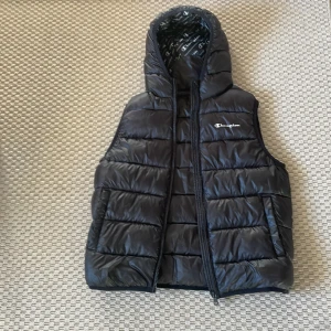 Svart dunväst med huva från Champion - Svart quiltad dunväst från Champion med huva och dragsko. Västen har dragkedja framtill och logga broderad på bröstet. Perfekt för lager-på-lager och streetwear. Insidan av huvan har tryckt Champion-mönster.