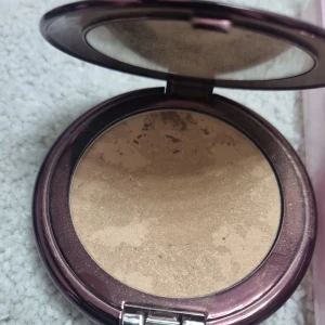 Kompakt bronzer i guldig nyans - Snygg bronzer i kompaktformat med spegel i locket. Färgen är en varm, guldig-brun ton med lätt skimmer som ger glow till ansiktet. Perfekt för att framhäva kindbenen eller ge solkysst look. Kommer i ett lila metallic-fodral.