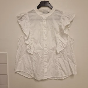 Vit blus med volang från ASOS DESIGN - Säljer en vit blus från ASOS DESIGN i bomull med knäppning framtill och volangdetaljer längs ärmhålen. Blusen är ärmlös och har en rak, lite loose passform. Perfekt för dig som gillar en söt och stilren look.