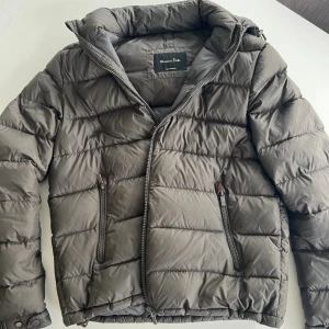 Pufferjacka från Massimo Dutti - Snygg olive/grå pufferjacka från Massimo Dutti med huva och två dragkedjeförsedda fickor. Jackan har quiltad design och stängs med dragkedja. Perfekt för kyliga dagar och ger en clean, modern look.