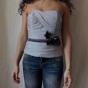 Ljusgrå bandeau tubtopp med blommor - Supersnygg ljusgrå bandeau tubtopp med draperad omlott framtill och dekorativa svarta blommor vid midjan. Toppen har en markerad midja med ett satinband och är helt axelbandslös. Perfekt för fest och finare tillfällen.