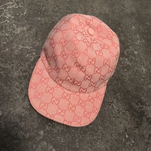 Rosa Gucci keps med GG-mönster - Säljer en rosa keps från Gucci med ikoniskt GG-mönster över hela kepsen. Justerbar rem baktill i ljusrosa läder och klassisk böjd skärm. Tillverkad i bomullstyg med exklusiv känsla och tydlig Gucci-label på insidan. Perfekt för dig som vill sticka ut med lyxig streetstyle.