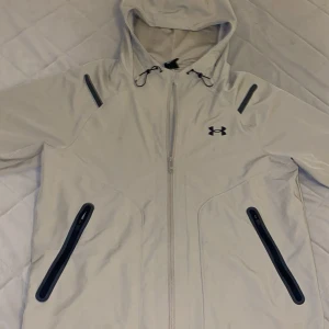 Beige vindjacka under armour  - Snygg beige vindjacka från Under Armour med huva och svarta detaljer. Jackan har dragkedja framtill, två fickor med svarta dragkedjor och justerbar snörning vid huvan. Tillverkad i lätt och slitstark polyester med meshfoder för extra komfort. Helt ny bara använd 6 gånger
