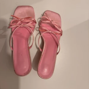 Rosa sandaler med rosett och klack - Säljer ett par ljusrosa sandaler med låg klack och fyrkantig tå. Skorna har tunna remmar i satin och två söta rosetter framtill. Insidan är också rosa och materialet har en glansig finish som ger en feminin vibe.
