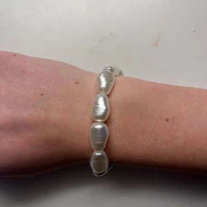 Armband med stora vita pärlor - Snyggt armband med stora, oregelbundna vita pärlor som ger en cool och unik look. Pärlorna har en skimrande yta och är trädda på elastisk tråd för enkel användning. Perfekt för dig som gillar statement-smycken och vill sticka ut lite extra.