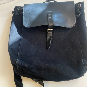 Svart ryggsäck från Sandqvist i canvas - Stilren svart ryggsäck från Sandqvist med lock i skinn och justerbara axelremmar. Väskan har snörning upptill, innerfack och detaljer i läder. Perfekt för dig som vill ha en klassisk och praktisk väska med snygga metallspännen.