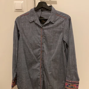 Blå skjorta med broderi från Limelight - Snygg blå skjorta från Limelight med färgglada broderade detaljer på ärmslut och en stor broderad patch på ryggen. Skjortan har knappar framtill, klassisk krage och lång ärm. Materialet känns som bomull och har ett diskret randigt mönster. Perfekt för dig som gillar unika plagg med coola detaljer.