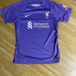 Lila Liverpool Nike fotbollströja - Säljer en lila Liverpool fotbollströja från Nike med Standard Chartered-tryck på bröstet och klubbmärke. Tröjan är kortärmad och tillverkad i ett lätt, ventilerande funktionsmaterial som passar perfekt för träning eller match.