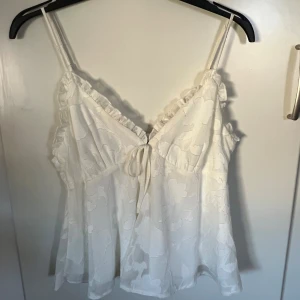 Vit volangtop från Gina Tricot - Supersöt vit linnetopp från Gina Tricot med volangdetaljer längs v-ringningen och tunna justerbara axelband. Toppen har ett diskret blommönster i tyget och knyt där fram. Den har två små defekter en lite fläck och en missfärgning vid kanten som man kan se på sista bilden 