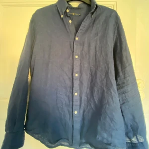 Blå linneskjorta från Ralph Lauren - Snygg blå skjorta från Ralph Lauren i 100% linne. Klassisk modell med knappar hela vägen, button down-krage och långa ärmar. Perfekt för varma sommar kvällar eller fester.