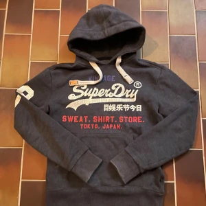 Mörkgrå Superdry hoodie med tryck - Snygg mörkgrå hoodie från Superdry med stort vitt och rött tryck på bröstet och siffran 28 i vitt på ena ärmen. Tröjan har huva med snörning och mjukt fleecefoder på insidan. Perfekt för dig som gillar streetwear och sportig stil.