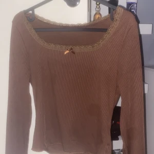 Brun ribbad långärmad topp med spets - Säljer en brun långärmad topp med ribbad struktur och söt spetskant runt halsringningen. Toppen har en liten rosett framtill som detalj och är figurnära i modellen. OBS! DE HÄR INGA FLÄCKAR DET ÄR BARA KAMERAN SOM ÄR BUGGAD (har bara haft på mig en gång