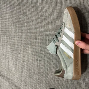 Adidas Gazelle i ljusgrå mocka - Säljer ett par Adidas Gazelle sneakers i ljusgrå mocka med vita klassiska ränder och gummisula. Skorna har snörning och diskreta bruna detaljer vid hälen. 