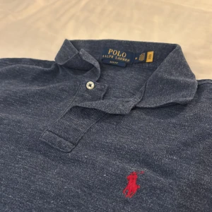 Blå pikétröja Polo Ralph Lauren M  - Marinblå pikétröja från Polo Ralph Lauren i M slim fit men passar även S. Tröjan har korta ärmar, krage med två knappar och den ikoniska röda broderade loggan på bröstet. 