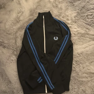 Svart track jacket från Fred Perry - Svart track jacket från Fred Perry med blå ränder längs ärmarna och vit dragkedja. Jackan har högt krage, ribbade muddar och Fred Perry-logga broderad på bröstet. Perfekt för en sportig och stilren look.