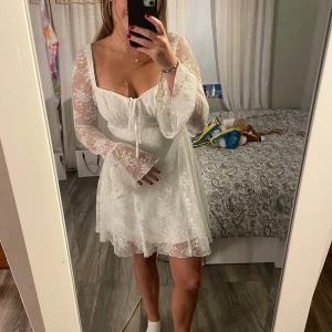 Studentklänning från Gina Tricot - Supersöt vit kortklänning från Gina Tricot med långärmar i spets och blommigt mönster. Klänningen har en urringning med snörning framtill och lätt utsvängda ärmslut. Perfekt för studenten💕