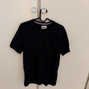 Svart t-shirt från Tommy Jeans - Svart t-shirt från Tommy Jeans i klassisk modell med rund hals och diskret logga på bröstet. T-shirten har korta ärmar och en snygg randig detalj på insidan av kragen. Tillverkad i mjuk bomull för skön känsla.