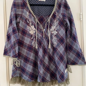 Rutig blus från Odd Molly med spets - Superfin rutig blus från Odd Molly i blått, rött och vitt med broderade blommor framtill. Blusen har v-ringning med knytband, spetsdetaljer vid ärmslut och nederkant samt vida trekvartsärmar. Perfekt för dig som gillar boho-vibbar och unika detaljer.        Mått  Över bröstet: ca 50 cm  Längd från axeln och ner: ca 75 cm   100% bomull   Kommer från ett djur och rökfritt hem. 