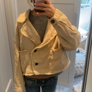 Beige croppad jacka - Säljer en beige croppad jacka från Zara med råa kanter och stora bruna knappar. Jackan har två fickor framtill, bred krage och är tillverkad i ett grovt bomullstyg. Perfekt för dig som gillar en chill och trendig look.