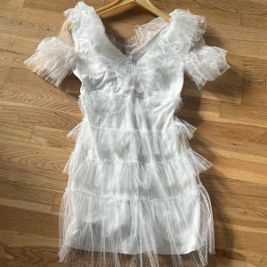 Vit kort klänning med volangtyll - Säljer min superfina studentklänning som är vit kort klänning med lager av transparent volangtyll och v-ringad hals och rygg. Klänningen har korta ärmar i tyll och dragkedja i ryggen. Materialet är en mix av polyester och tyll som ger en fluffig och unik look. Perfekt för dig som vill sticka ut med en lekfull och trendig stil.