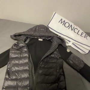 Svart dunväst med huva från Moncler - Helt ny Snygg svart dunväst från Moncler med glansig finish och quiltad design. Västen har huva, dragkedja framtill och Moncler-logga på ärmen. Perfekt för lager-på-lager och streetstyle. Materialet är dun och polyester.