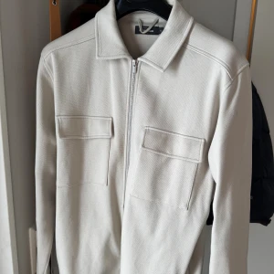 Beige overshirt från Dressmann M - Stilren beige overshirt från Dressmann i storlek M. Jackan har två stora bröstfickor med lock, klassisk krage och dragkedja framtill. Materialet är bomull och ger en clean och modern look. Perfekt för lager-på-lager och enkel att matcha. Finns lite fläckar.