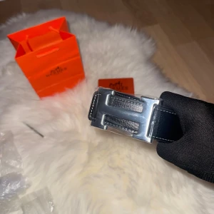Svart H skärp med H-spänne - Säljer ett stilrent svart H bälte i skinn med klassiskt H-spänne i silverfärgad metall. Bältet har en slät, elegant form och vit kontrastsöm längs kanterna. Perfekt accessoar för att lyfta din outfit med lyxig känsla.box medkommer ej håltagare medkommer