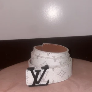 Vit bälte  - Snyggt vitt bälte med klassiskt monogrammönster och stor svart spänne. Bältet är gjort i skinn och har en stilren look som lyfter vilken outfit som helst.